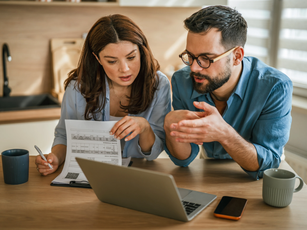 Man and woman looking at financial documents and a laptop computer in a well-lit kitchen.