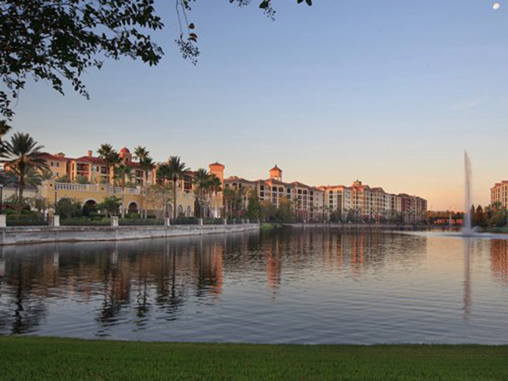 Scenic exterior view of Tuscany Village resort buildings reflecting across a calm lake with a fountain.