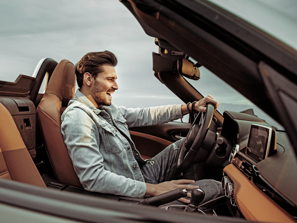 Smiling man in a denim jacket driving an open-top convertible along a scenic, winding mountain road.