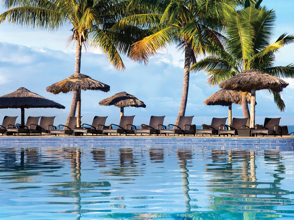 An outdoor pool reflecting palm trees and thatched umbrellas with lounge chairs at the water's edge at WorldMark Fiji.