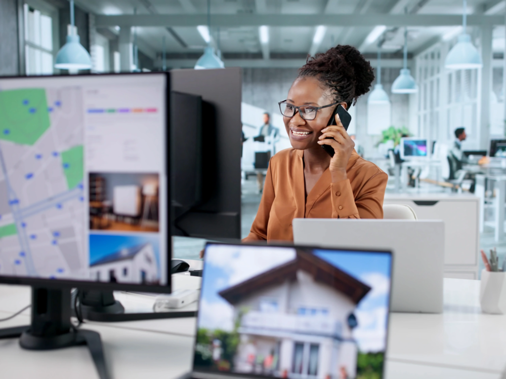 Smiling female real estate professional talking on a smartphone in a bright, modern office, with property listings and a map displayed on her computer monitors.