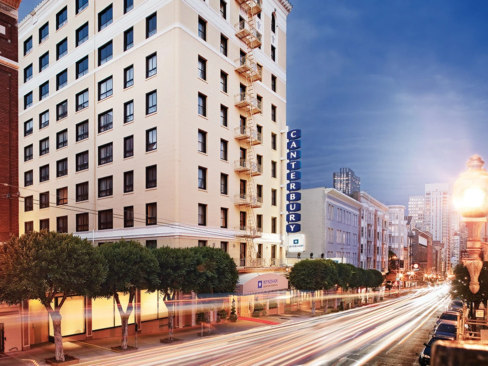 The Canterbury Hotel with a lit sign, trees, and city street in evening light.