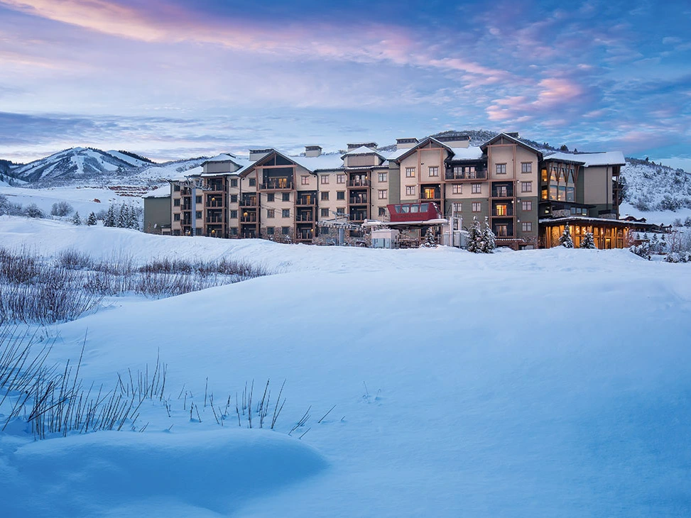 A snow-covered building surrounded by mountains during twilight, with soft pink and blue skies.