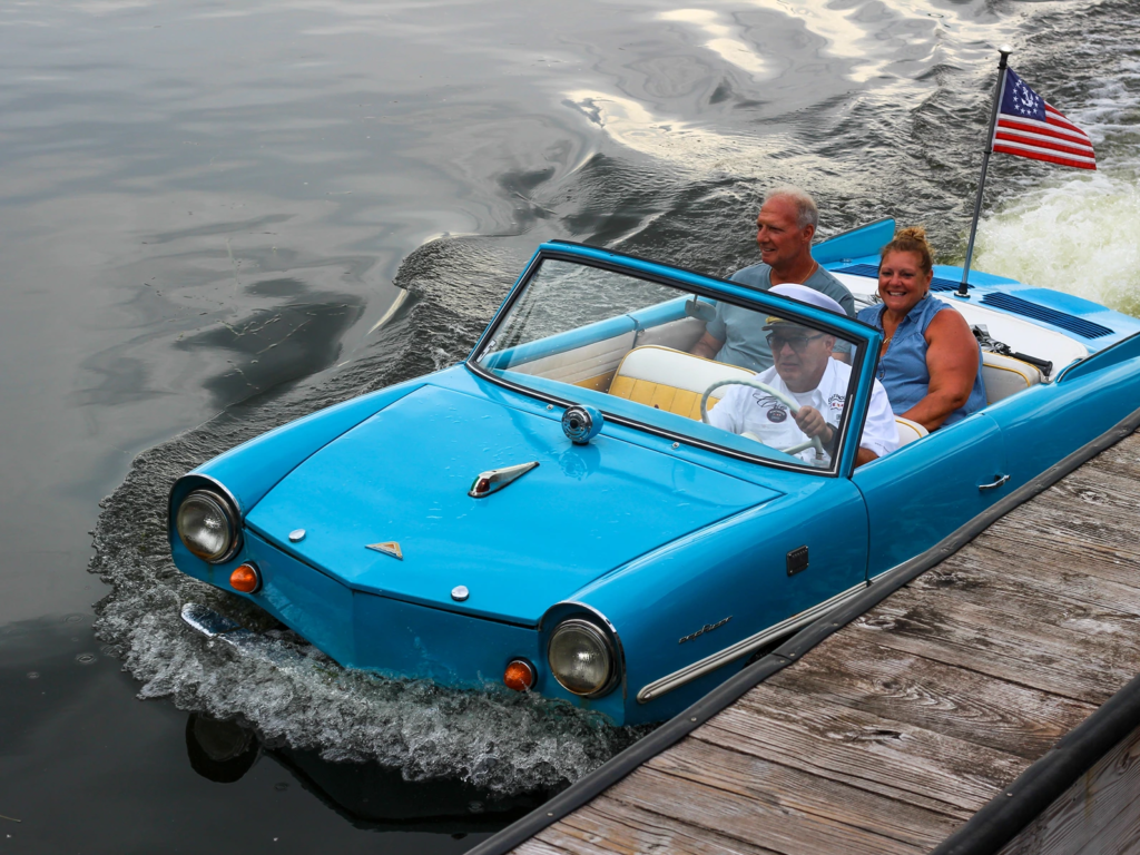 A blue convertible Amphicar with three people, docking at Disney Springs.