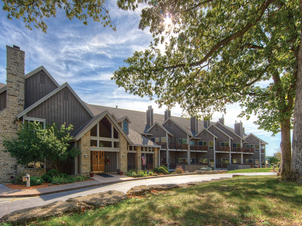 A large lodge-style building with wooden siding and stone details, surrounded by trees and grassy area.