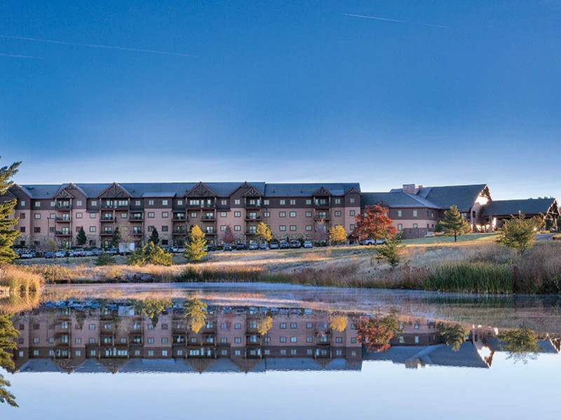 Sell Wyndham Points: A hotel building reflects in a calm lake, surrounded by trees and grass in a clear sky.