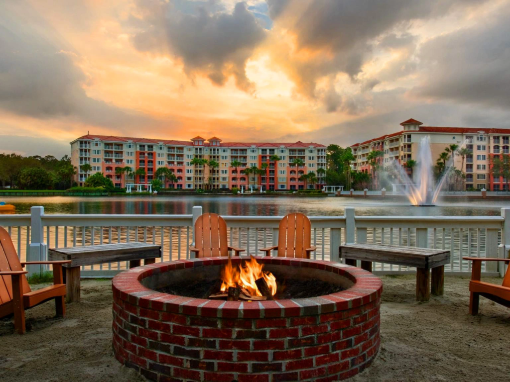 A cozy fire pit at Grande Vista with flames, surrounded by wooden chairs near a lake at sunset.
