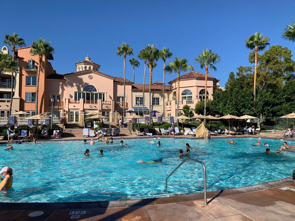 A sunny pool area at Newport Coast Villas with people swimming, palm trees, and lounge chairs.