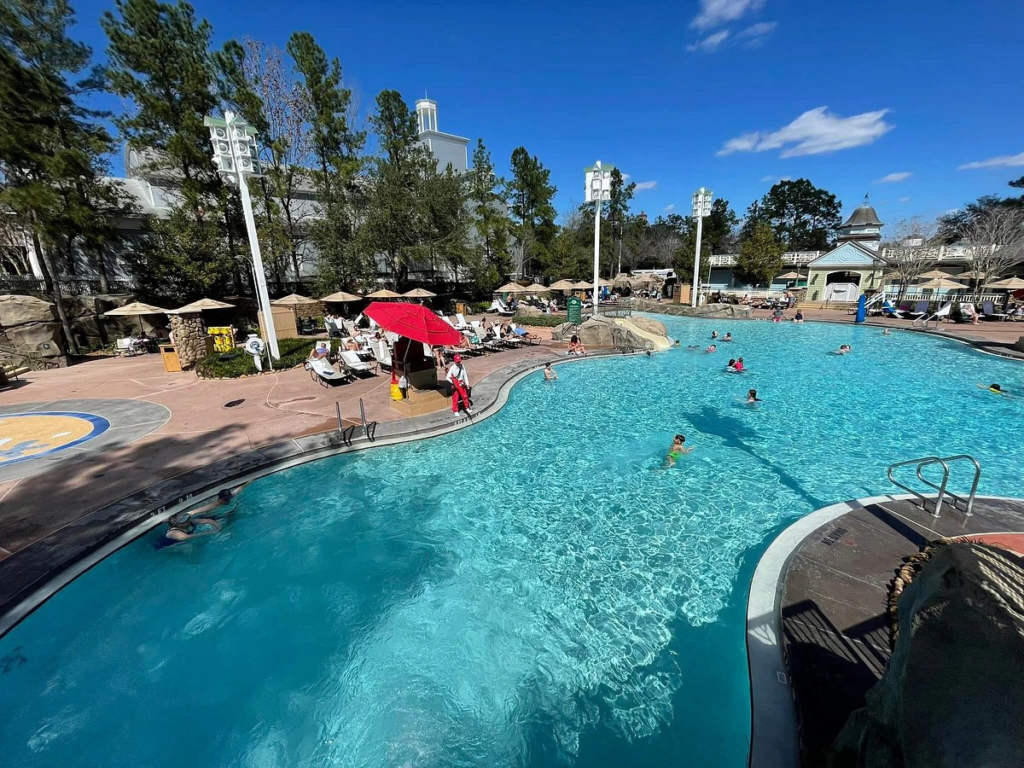 A sunny pool area at Saratoga Springs with people swimming, lounge chairs, and an umbrella. Trees and buildings in the background. Saratoga Spring DVC Resale
