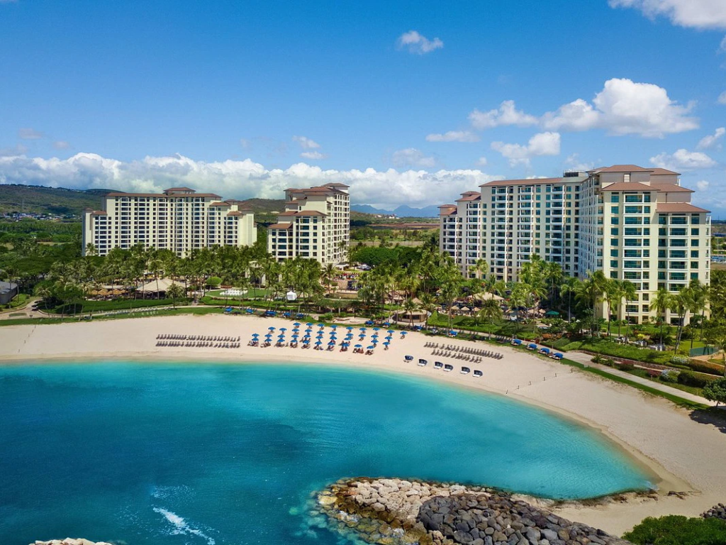 Aerial view of Marriott Ko Olina Beach Resort with tall buildings, palm trees, and blue umbrellas on the sand.