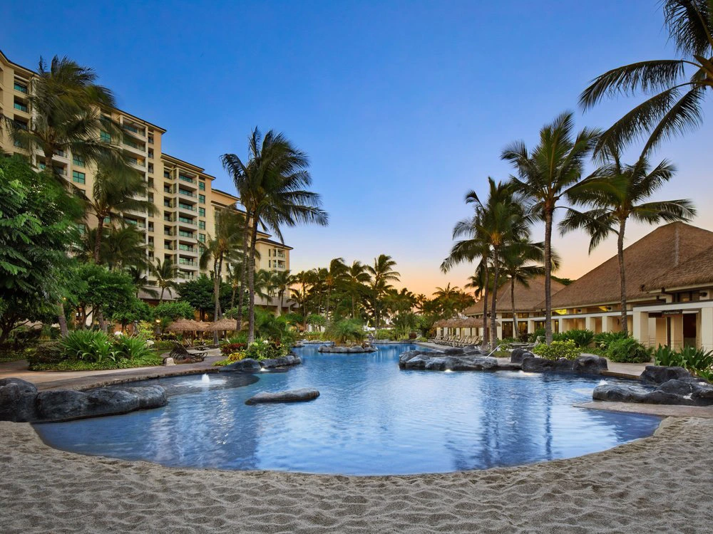 A tranquil resort pool at Marriott Ko Olina surrounded by palm trees, rocks, and tropical plants at sunset.