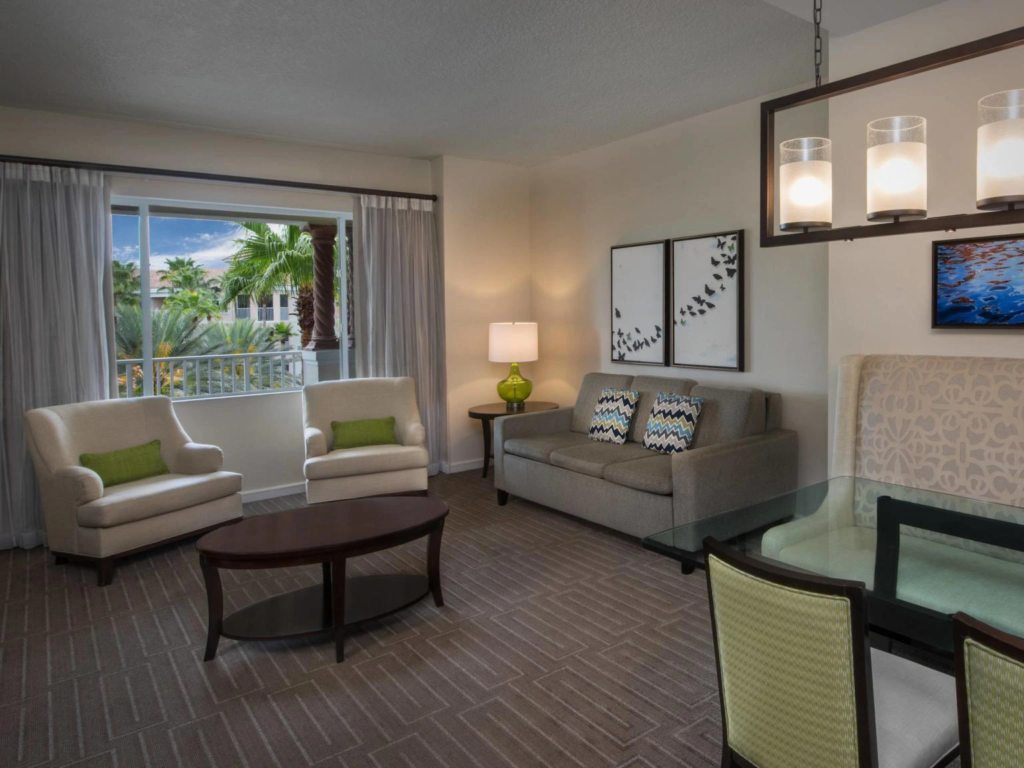 A cozy living room at Marriott's Grande Vista with two armchairs, a sofa, a dining table, and big windows showcasing palm trees outside.