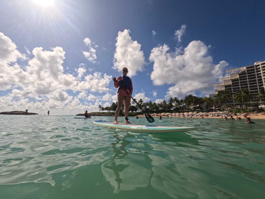 A woman stand-up paddleboarding on clear, sunny turquoise water at the aulani disney vacation club villas beach.
