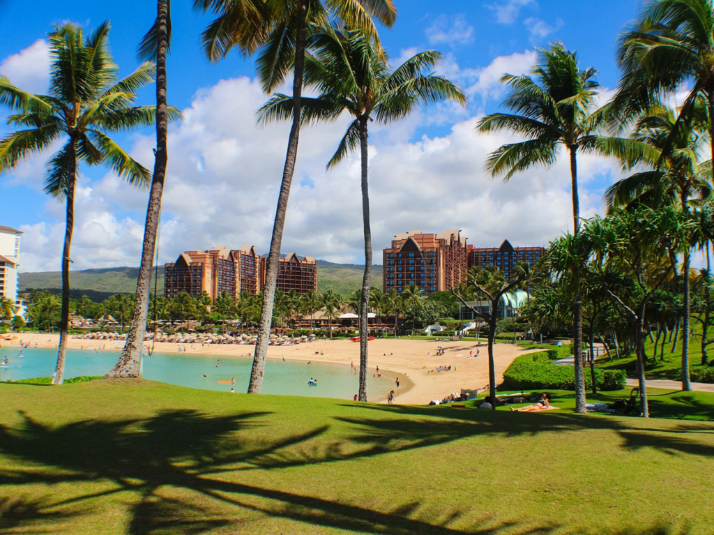 View of Aulani resort towers across a white sand beach lagoon framed by tall palm trees in Ko Olina, Hawaii.