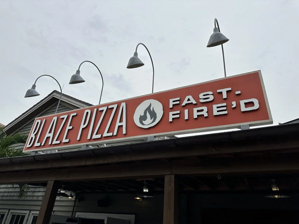 An orange sign for a fast-casual pizza restaurant mounted below curved vintage light fixtures on a wooden roof overhang.