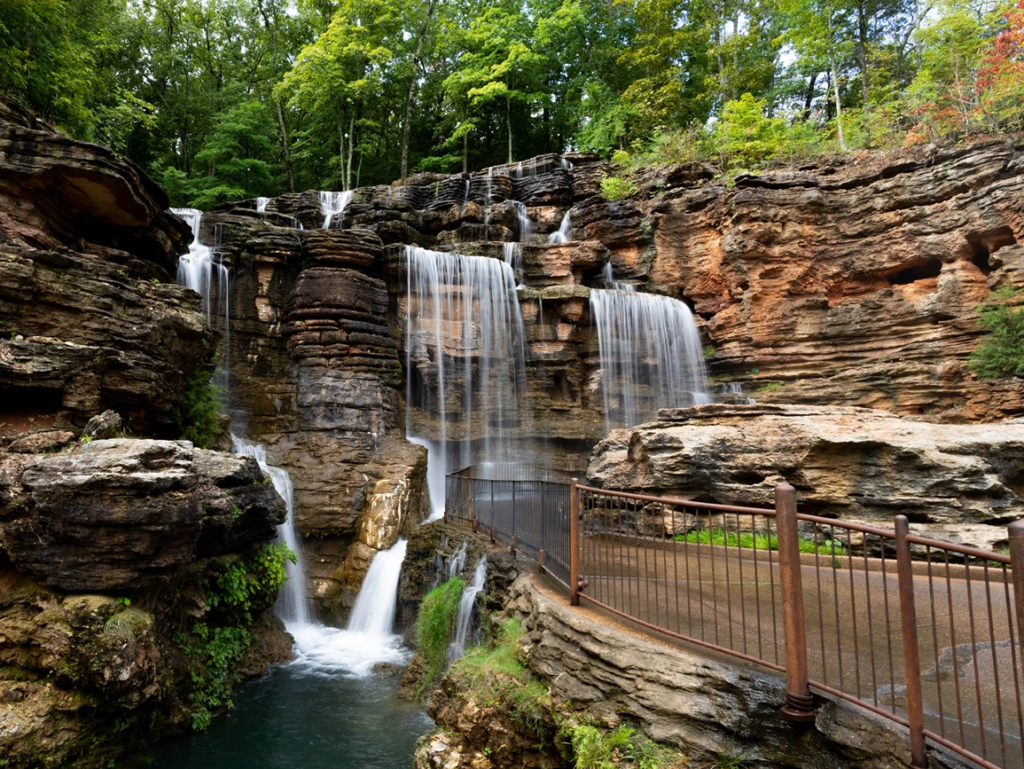 A scenic multi-tiered waterfall cascading over rocky ledges next to a paved walkway.