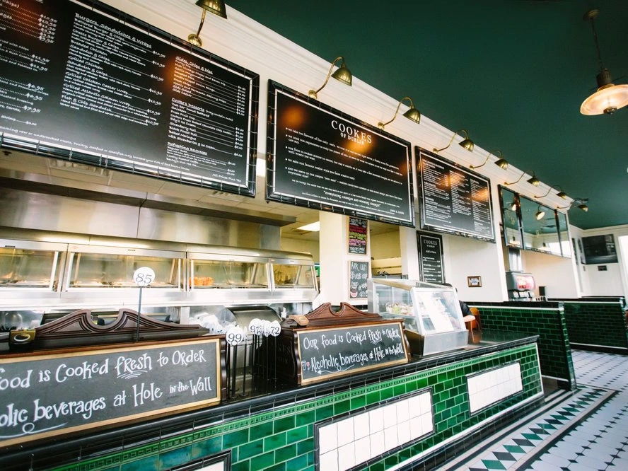 Bright restaurant service interior featuring glossy green tiled ordering counters and hanging framed menu boards.