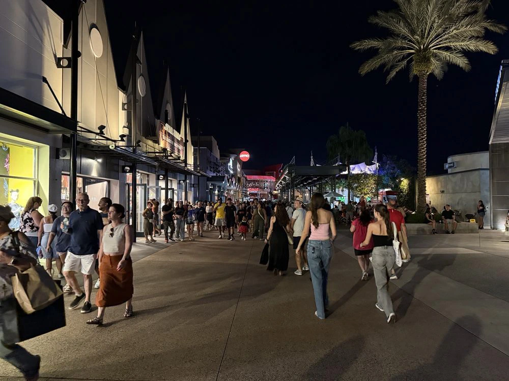 Visitors stroll along a lively outdoor shopping promenade at Disney Springs at night under illuminated storefronts.