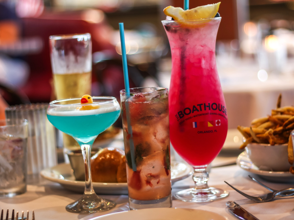 Three colorful craft cocktails, including a bright blue drink topped with a rubber duck, sit on a restaurant dining table.
