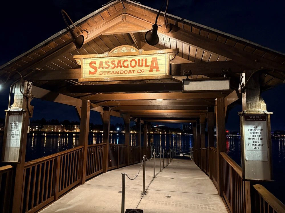 A covered wooden ferry dock at Disney Springs at night, with calm water reflecting illuminated buildings across the lake.