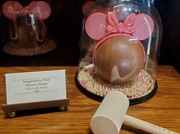 A chocolate Minnie Mouse sphere treat with pink ears sits under a glass dome beside a small wooden serving mallet.