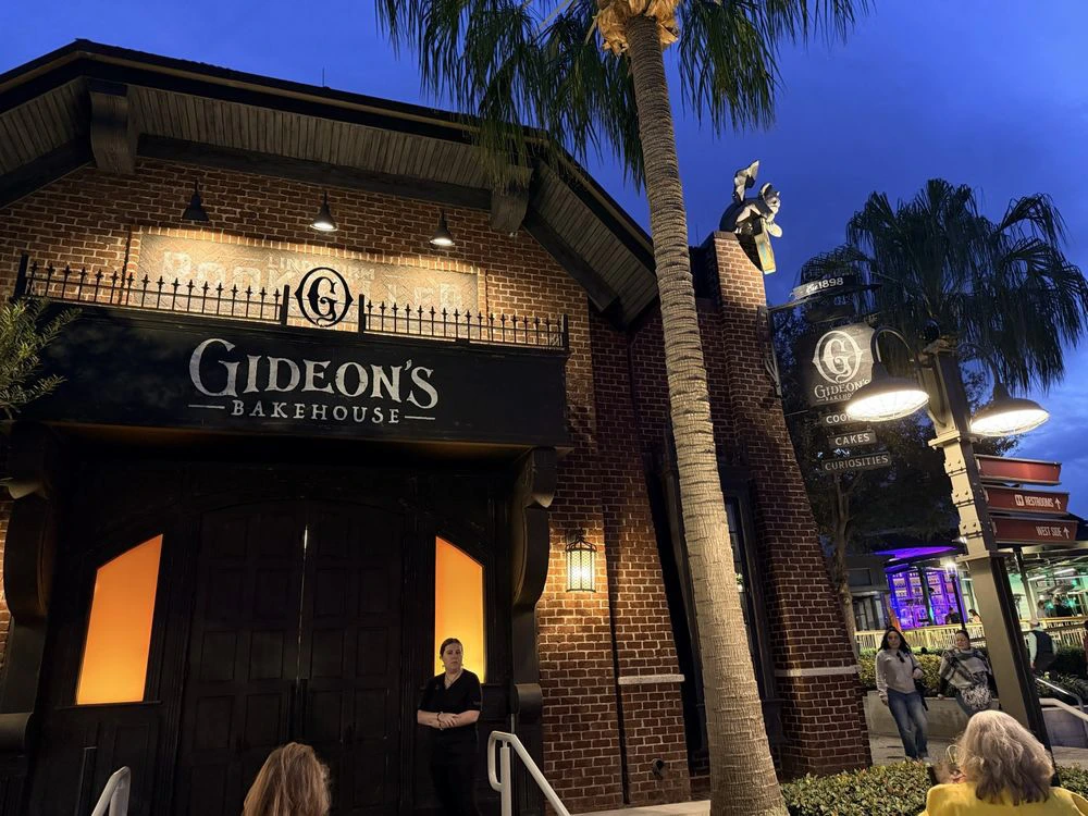 Patrons stand outside the glowing brick exterior and dark wooden doors of Gideon's Bakehouse in Disney Springs at dusk.