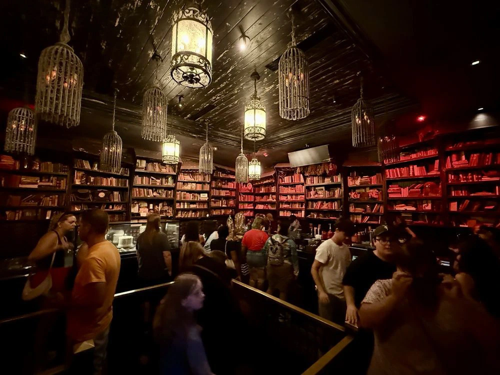 A dimly lit, crowded bakery interior lined with tall bookshelves and illuminated by hanging glowing birdcage lanterns.