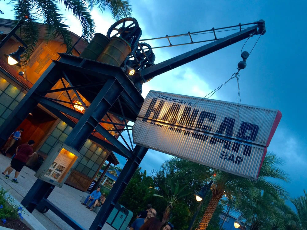 An industrial waterfront bar exterior featuring a large vintage crane suspending a corrugated metal entrance sign at dusk.