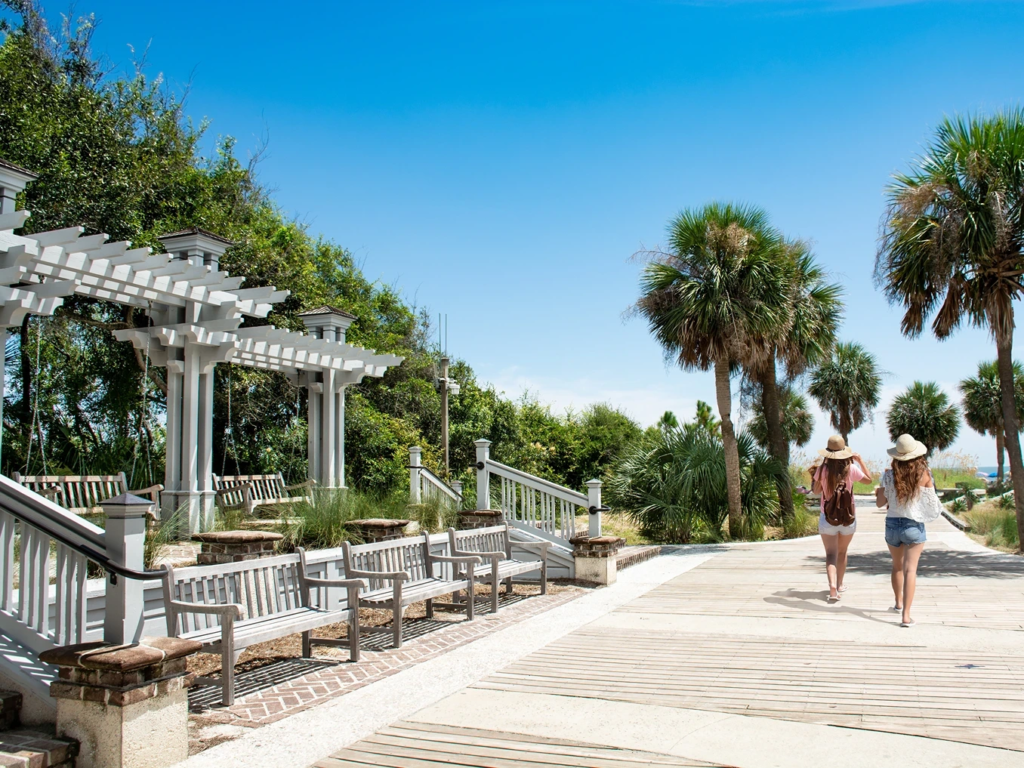 Two people walking down a sunny wooden beach boardwalk lined with palm trees in Hilton Head.