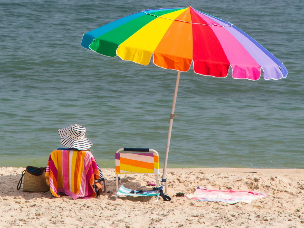 A person sitting on a sandy beach under a large, vibrant rainbow-colored umbrella.