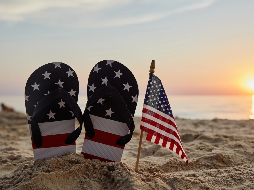 American flag themed flip-flops and a small US flag planted in the sand at sunset.