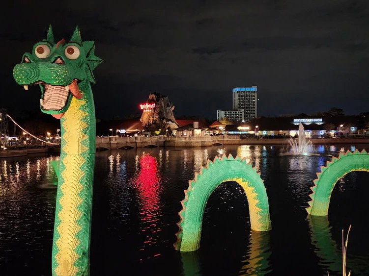 A giant green sea serpent sculpture made of toy bricks rises from the dark lake waters at Disney Springs at night.