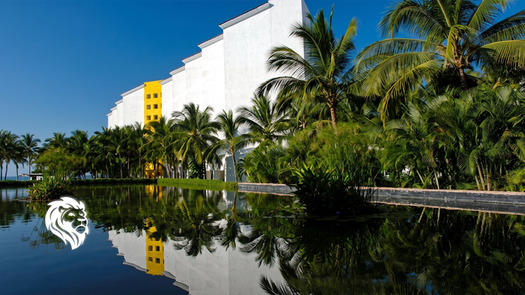 White stepped architecture of Mayan Palace reflected in a tranquil water feature surrounded by palms.