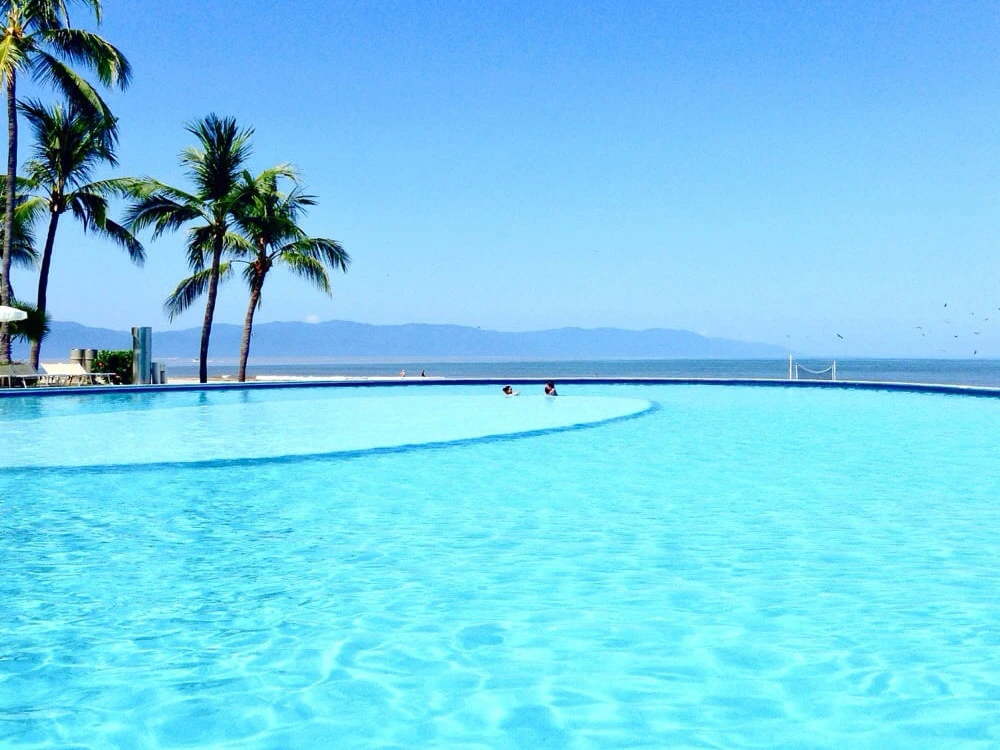 Expansive Mayan Palace infinity pool overlooking the blue ocean and distant mountains under a sunny sky.