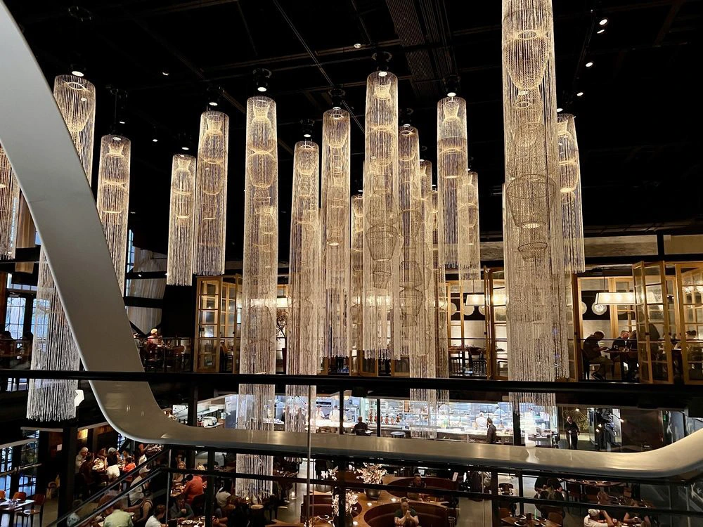 Diners enjoy a meal inside a spacious, multi-level restaurant featuring dramatic long glowing cylindrical chandeliers.