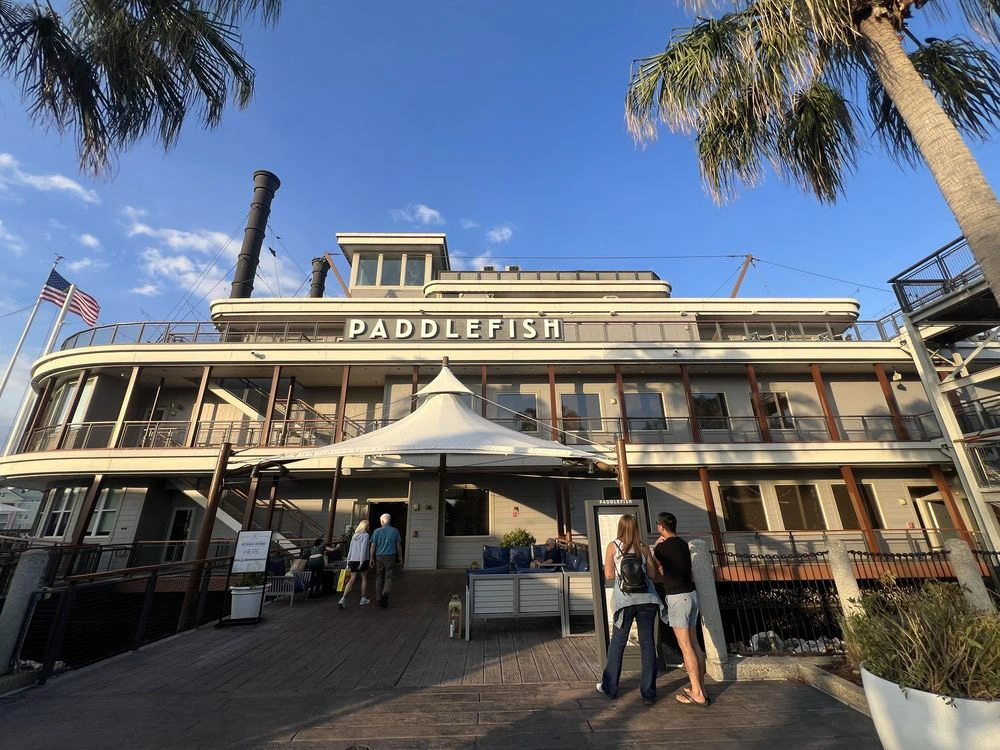 Guests walk along a wooden boardwalk toward a large stationary steamboat restaurant docked on the waterfront.