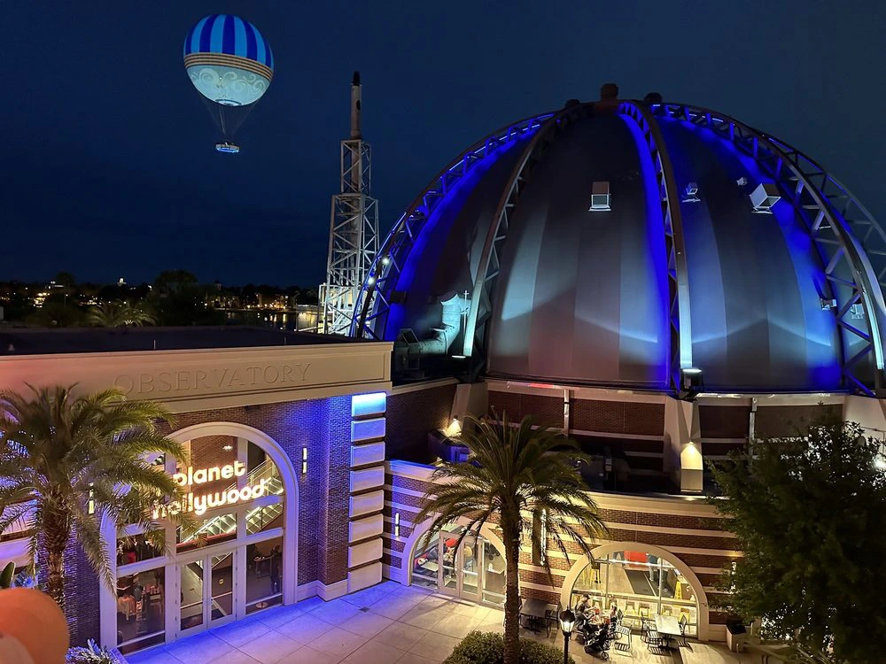 A glowing blue observatory dome stands at Disney Springs below an illuminated tethered hot air balloon in the night sky.