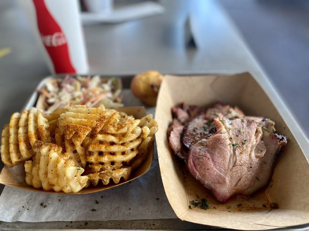 A barbecue platter featuring sliced smoked meat topped with herbs, seasoned waffle fries, coleslaw, and a small bread roll.