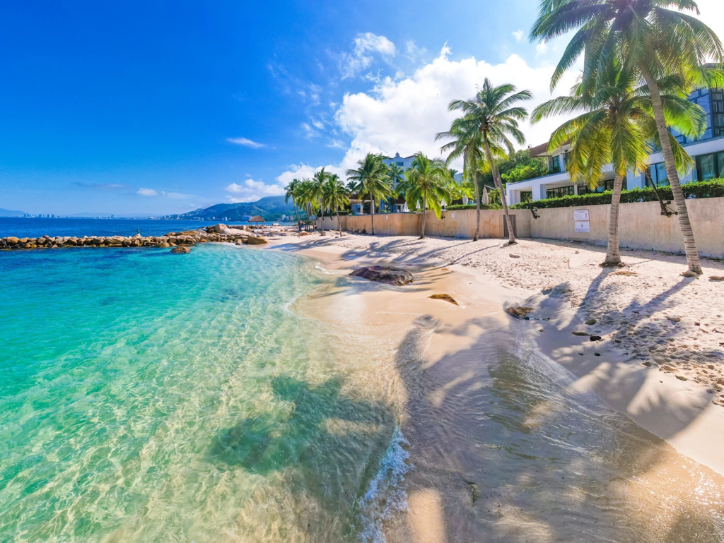 Clear turquoise ocean waves gently washing onto a sunny Puerto Vallarta beach lined with palm trees.