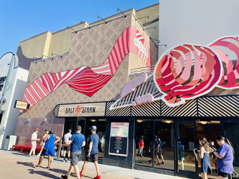 Visitors walk past the sculpted pink and red ice cream cone facade of Salt & Straw at Disney Springs.