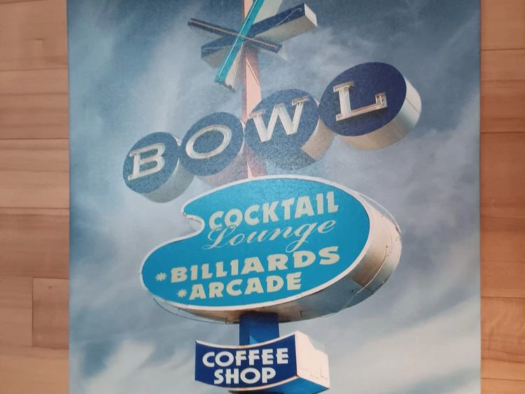 Retro wall sign advertising a bowl, cocktail lounge, billiards arcade, and coffee shop against a cloudy sky.