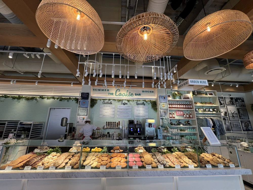 A brightly lit bakery counter displaying rows of large artisan cookies under hanging woven basket pendant lights.