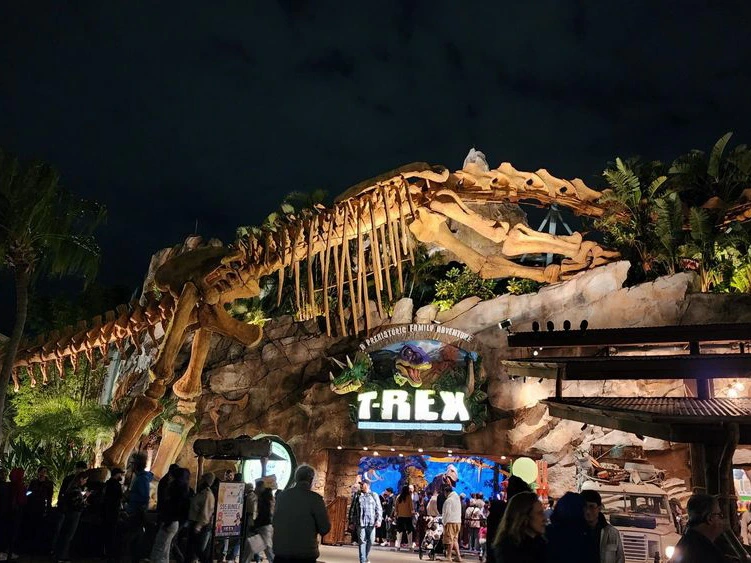 A massive dinosaur skeleton towers over the rocky entrance archway of the T-REX Cafe at Disney Springs at night.