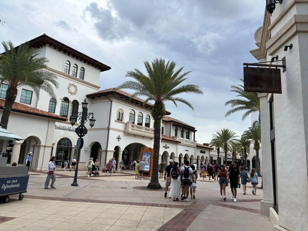 Visitors explore a wide outdoor plaza at Disney Springs lined with Spanish-style white buildings and a central palm tree.