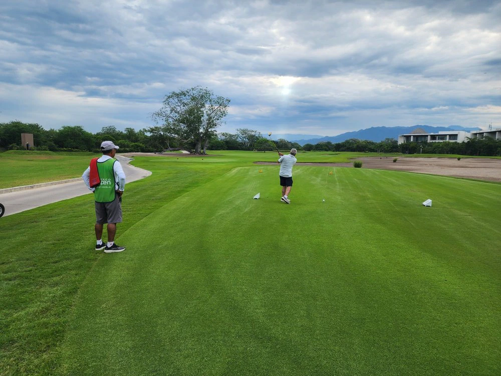 Golfer swinging on a lush green Vidanta golf course fairway as a caddie watches, with mountains under a cloudy sky.