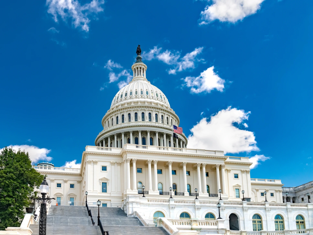 The white dome and grand steps of the United States Capitol building in Washington, D.C. under a bright blue sky.