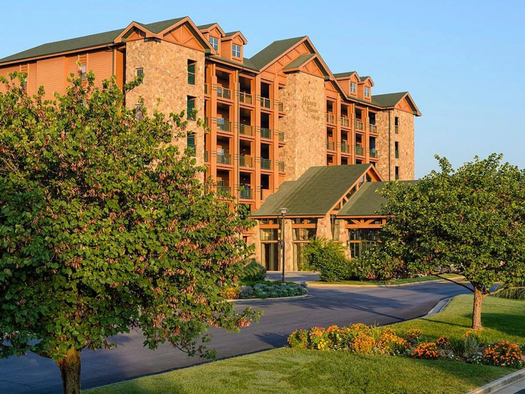 Multistory stone and wood lodge building surrounded by lush green trees and flowerbeds.