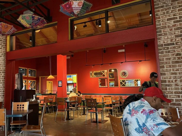 Diners eat at tables inside a vibrant restaurant with bright red walls and colorful patterned umbrellas on the ceiling.