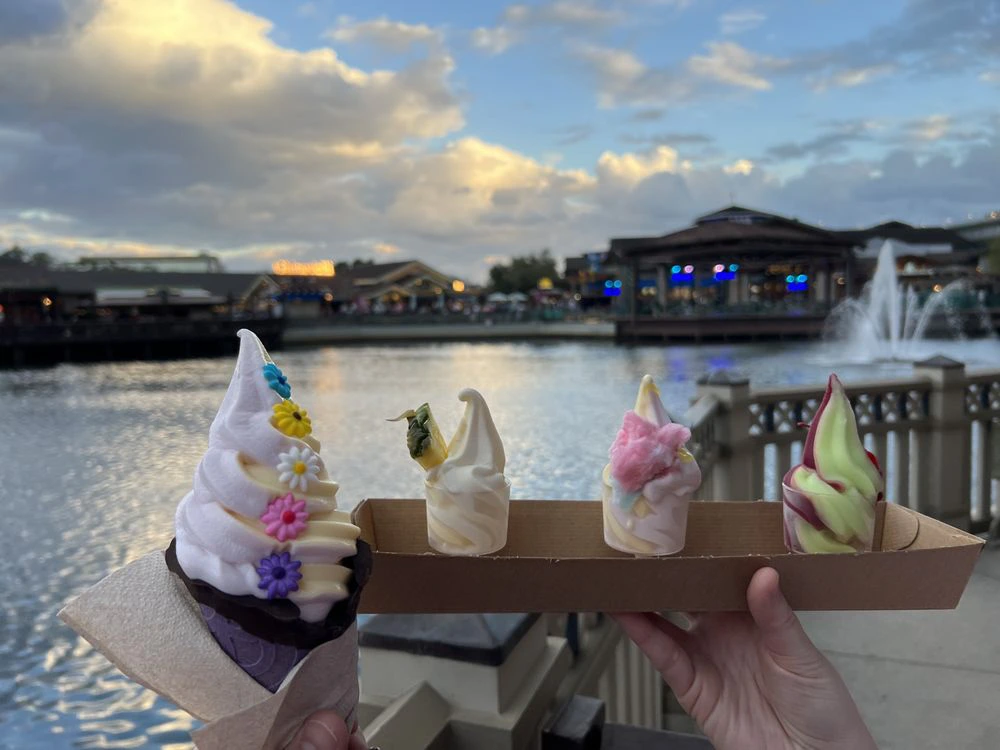 Four colorful soft-serve ice cream treats held up against a scenic waterfront background at sunset in Disney Springs.