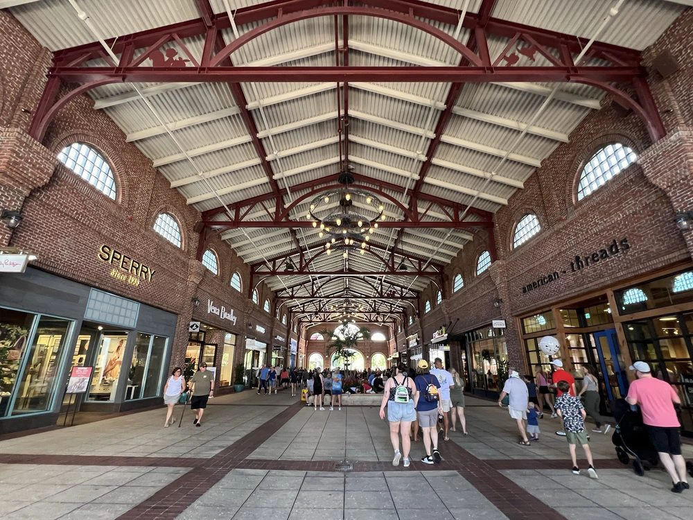 Visitors stroll through a grand covered brick shopping promenade featuring red steel roof trusses and iron chandeliers.